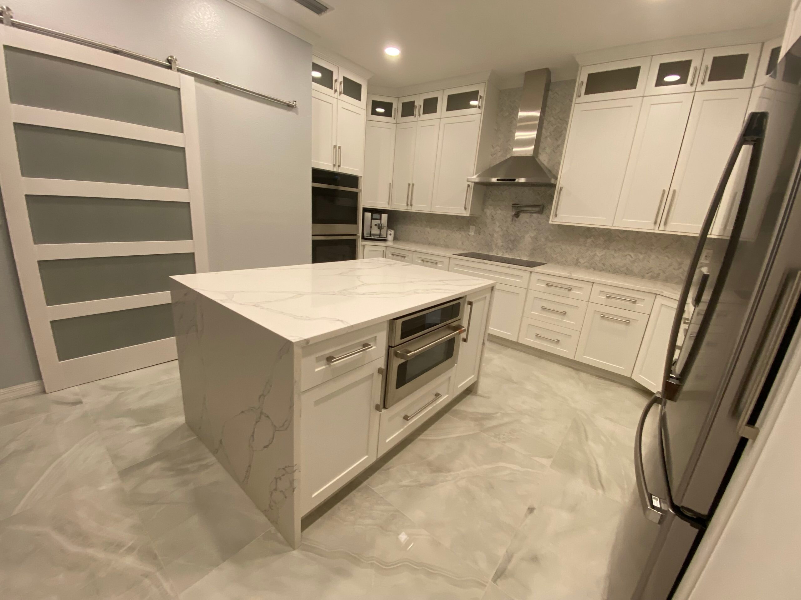 White shaker kitchen with marble waterfall island barn door double wall ovens and marble tile floors by 1 Stop Cabinets