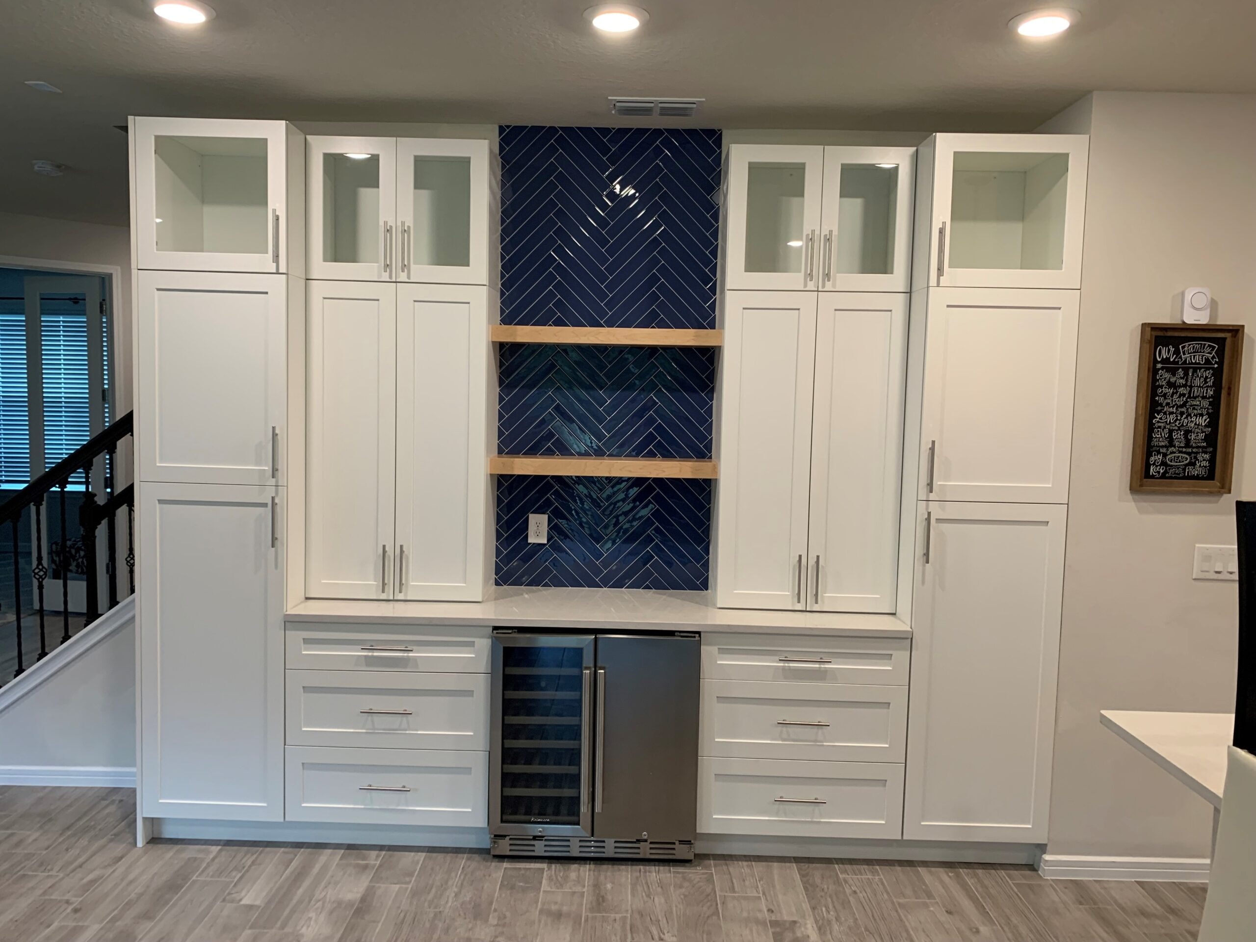 White shaker built-in bar wall with navy herringbone tile backsplash wood shelves and wine cooler by 1 Stop Cabinets