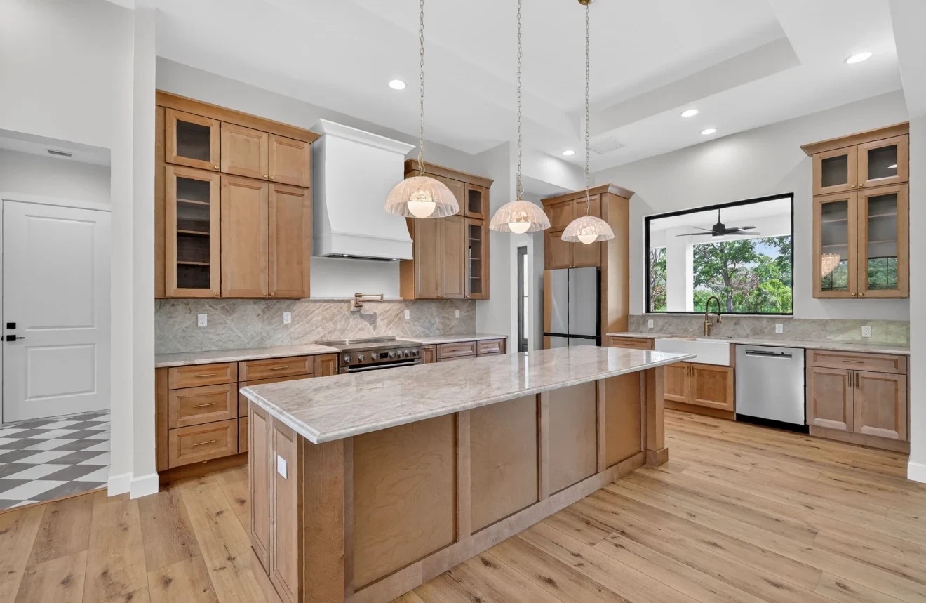 Natural maple raised panel kitchen with white custom range hood marble quartz island glass-front upper cabinets and farmhouse sink on hardwood floors by 1 Stop Cabinets