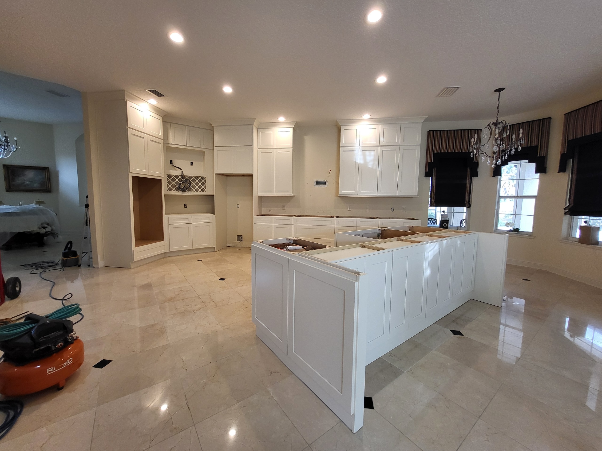 Large white shaker kitchen under construction with oversized island on marble tile floors by 1 Stop Cabinets